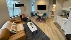 Ein modernes Wohnzimmer mit einer Couch, einem Couchtisch und einem Fernseher. Es gibt eine Essecke mit Tisch und Stühlen sowie eine Küche mit Schränken und einem Herd.