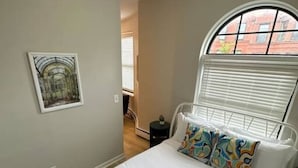 Ein Schlafzimmer mit Bett, einem Fenster mit Jalousien, einem Bild an der Wand und einem kleinen Tisch.