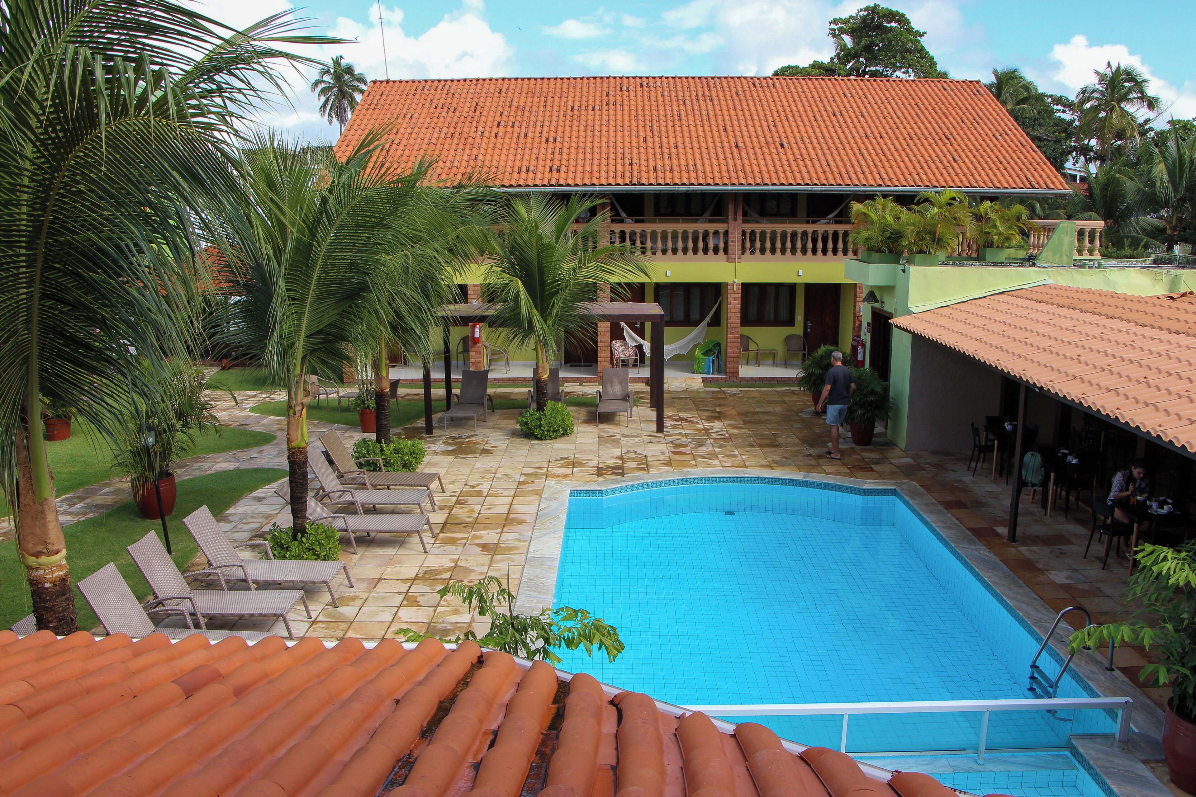 A swimming pool with lounge chairs, a covered patio, and a building with a tiled roof.