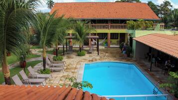 A swimming pool with lounge chairs, a covered patio, and a building with a tiled roof.