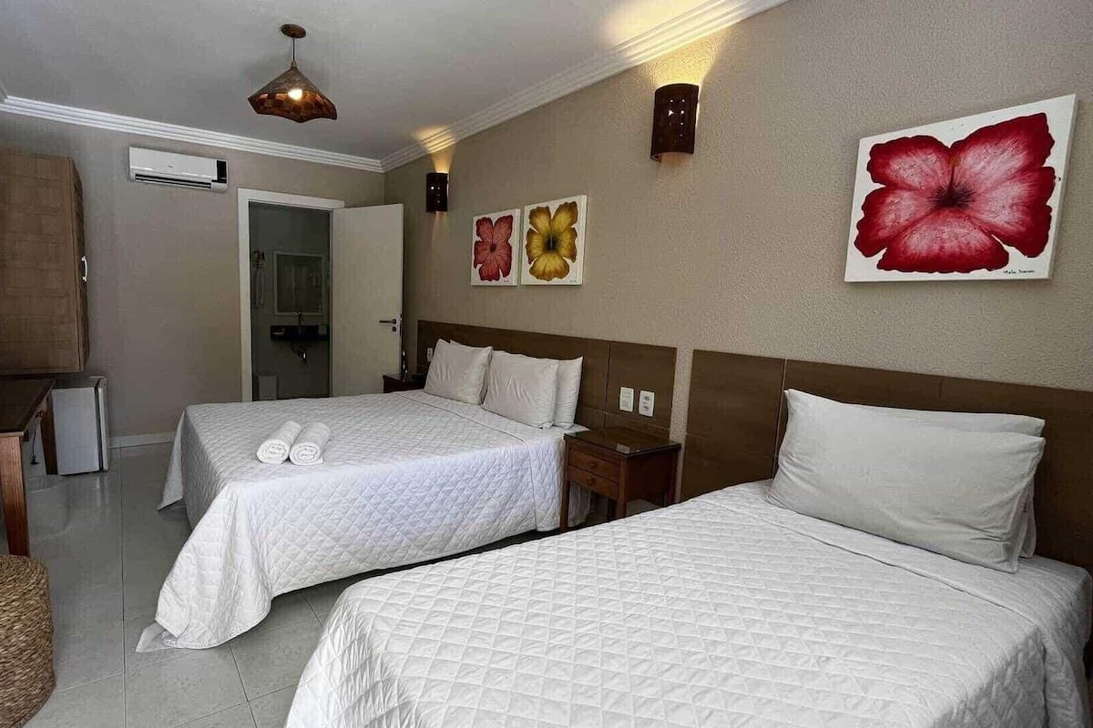 Habitació d'hotel amb dos llits, una tauleta de nit de fusta, una taula petita i una cistella teixida. Hi ha llums penjades a la paret i un quadre gran amb un floreig.