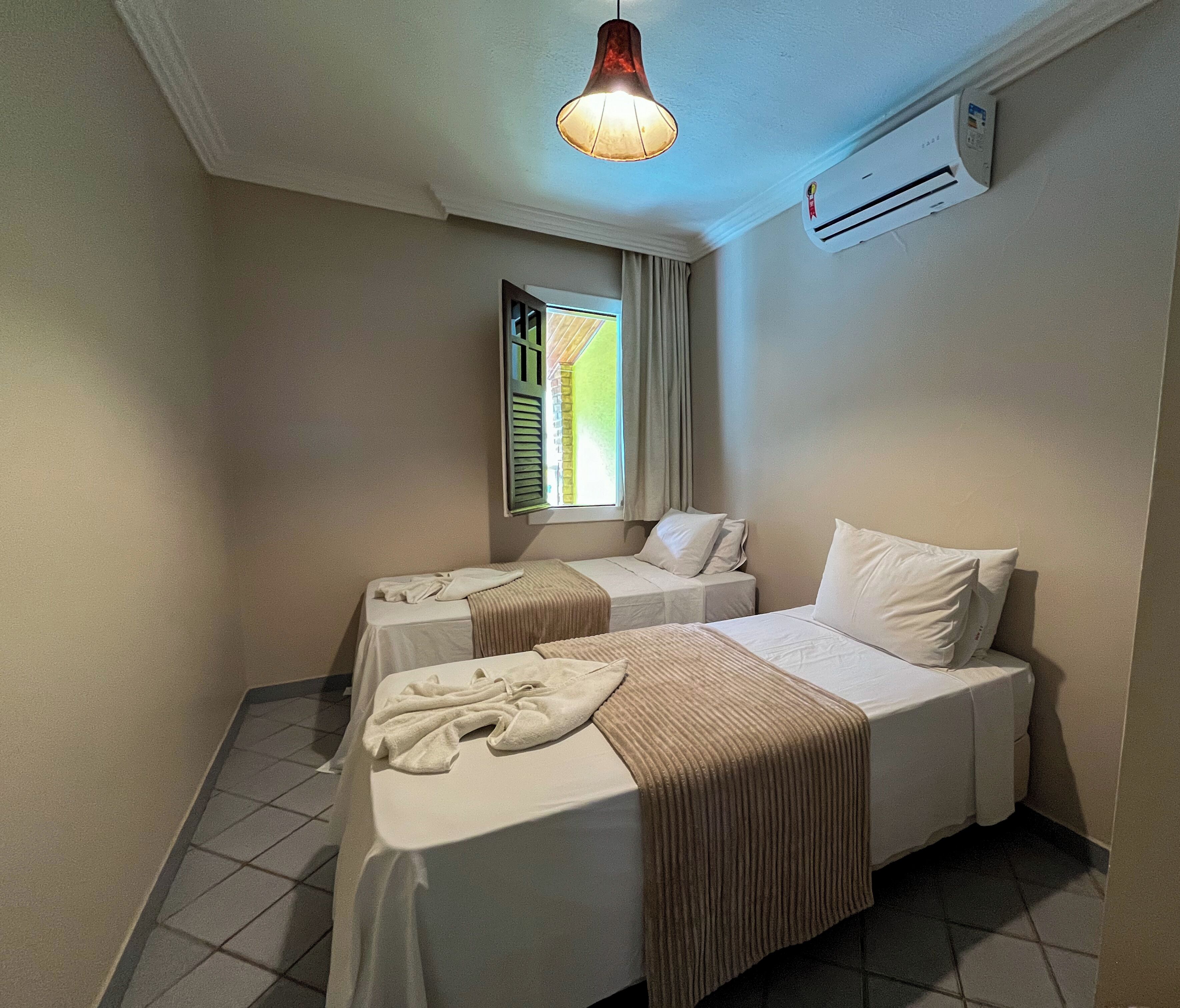 Quarto de hotel com duas camas, uma janela com cortinas, um lustre e um ar condicionado.