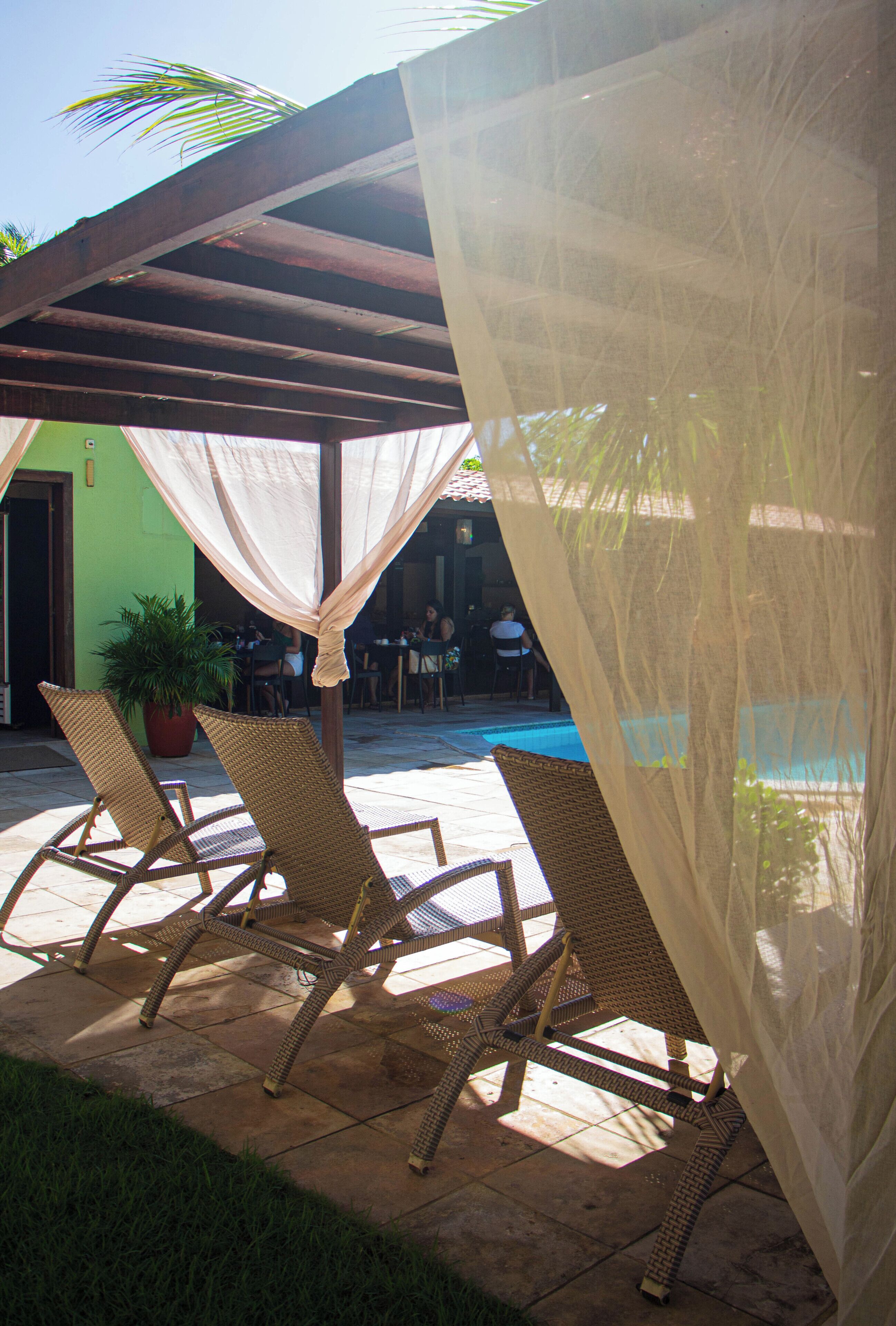 A covered outdoor seating area with wicker chairs, a potted plant, and a pool in the background.