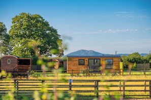 A wooden cabin with a large window and a door, surrounded by a fenced area with a tree and a mountain in the background. - Ockeridge Rural Retreats (Worcester)
