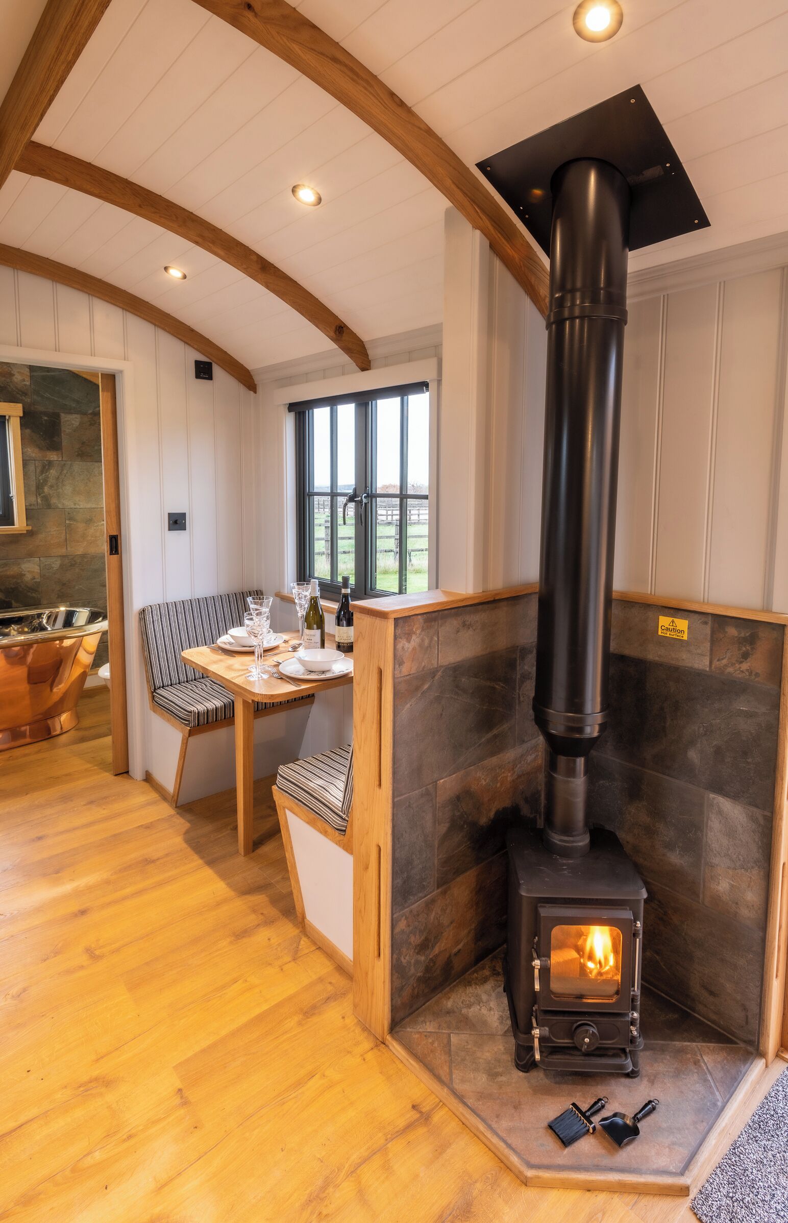 A cozy room with a wooden ceiling, a stone fireplace, a dining table with chairs, and a view of the outdoors through a window.