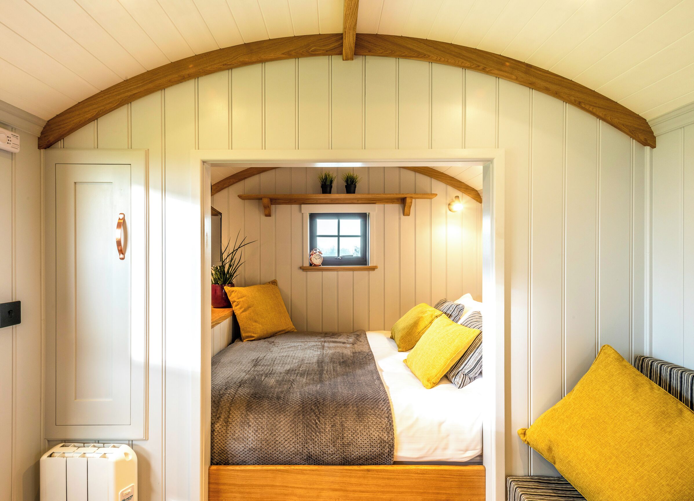 Ein gemütliches Schlafzimmer mit Holzbalkendecke, einem Bett mit grau-gelber Decke, einem kleinen Fenster und einem Regal mit Pflanzen.