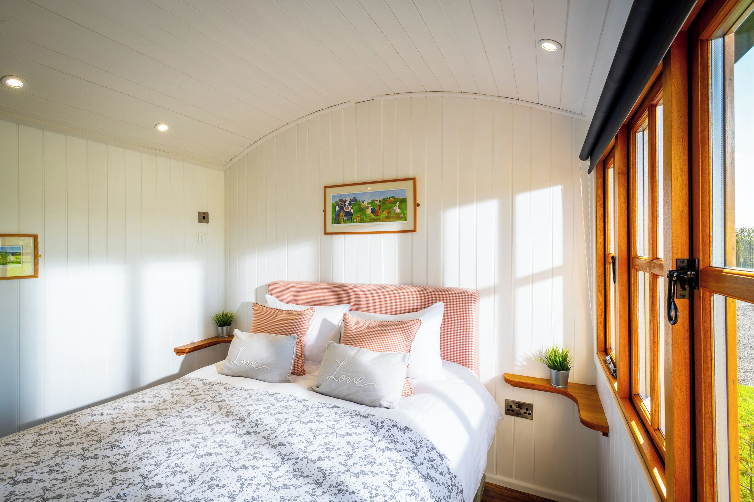 Une chambre à coucher avec un lit, des oreillers, un tableau encadré, une fenêtre et une étagère avec une plante.