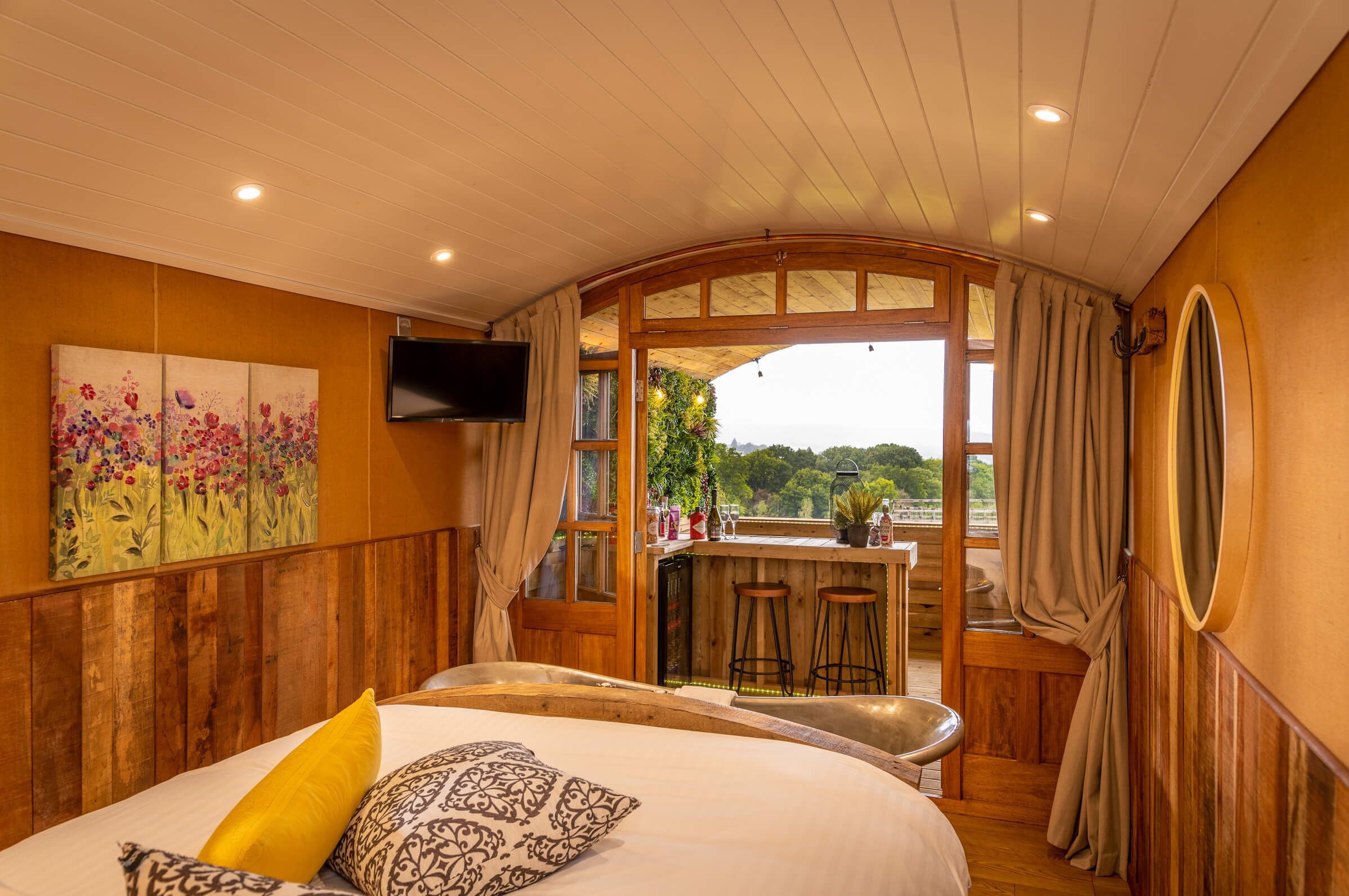 A bedroom with a large bed, a wooden headboard, a painting, a TV, and a balcony with a bar area.