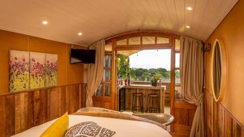 A bedroom with a large bed, a wooden headboard, a painting, a TV, and a balcony with a bar area.