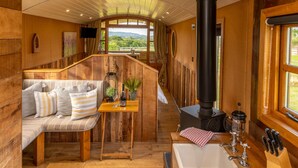 L'interno accogliente di una baita con una panca di legno, un tavolo con una bottiglia e dei bicchieri, una stufa nera e una finestra con vista sul verde.