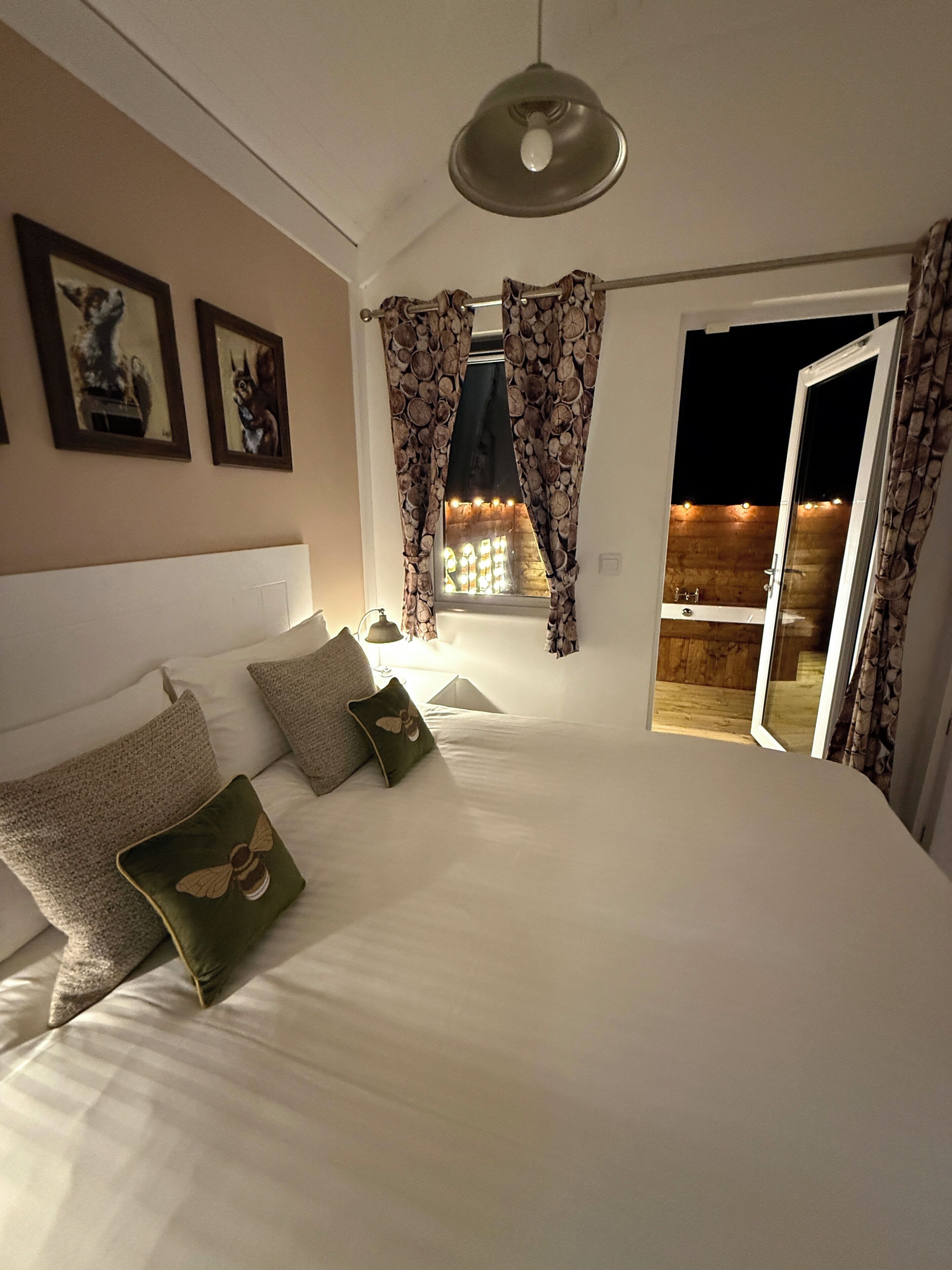Une chambre avec un grand lit, un luminaire au plafond et une vue sur une terrasse en bois, visible à travers une fenêtre ornée de rideaux.