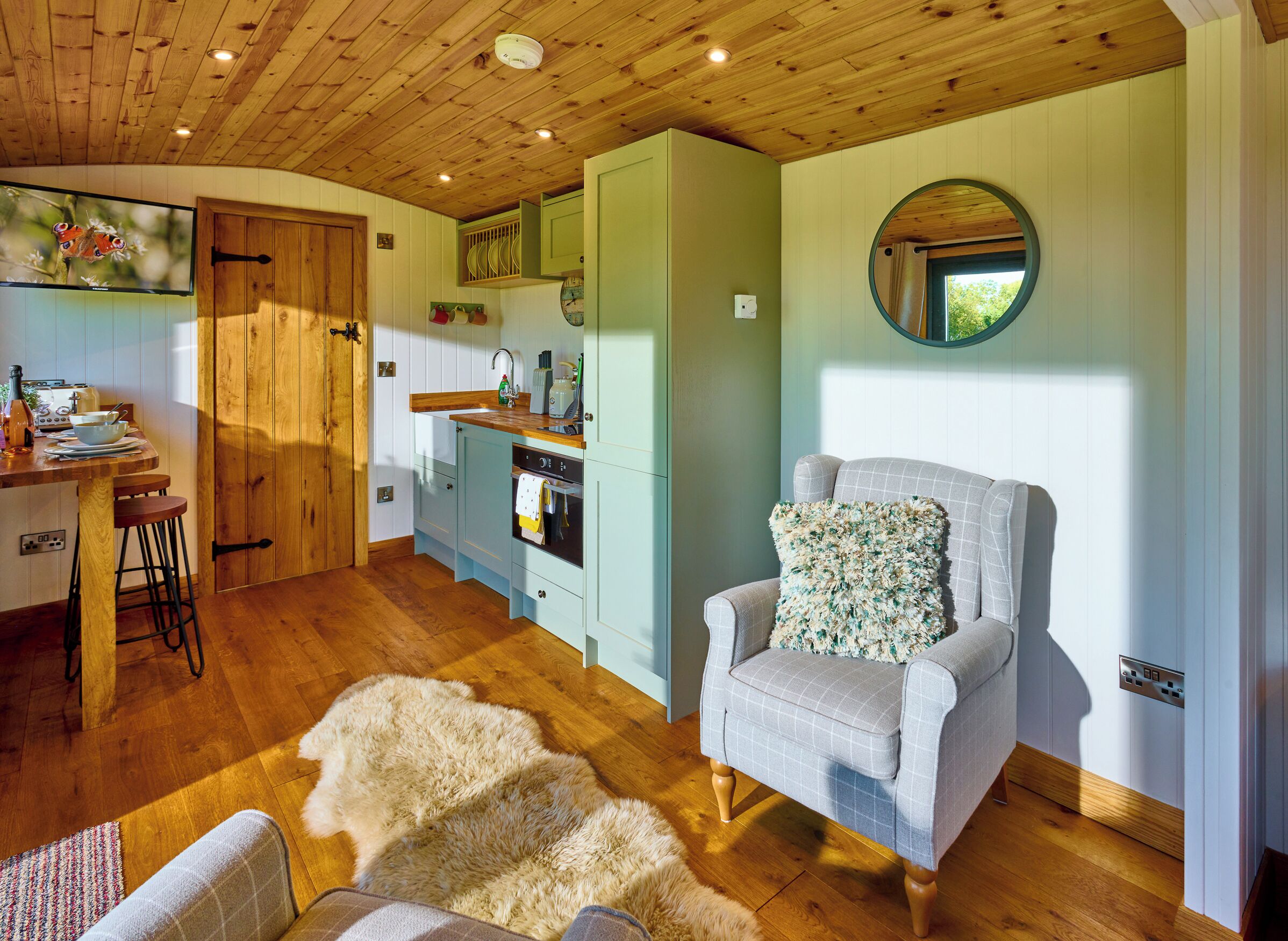 A cozy kitchen with wooden cabinets, a dining area with a bar stool, and a comfortable armchair with a floral pillow.