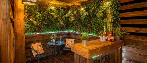 Een houten bar met een bord waarop 'GARDEN BAR' staat, omringd door houten wanden en een weelderige groene wand met planten en lichtslingers.