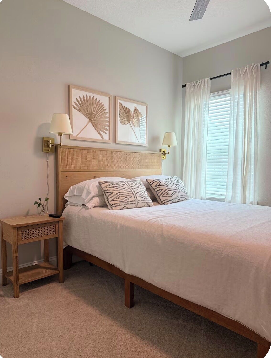 Un dormitorio con una cama de madera, dos lámparas fijadas a la pared, una mesita de noche con una planta y dos obras de arte con hojas enmarcadas.