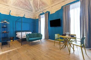 Ein Zimmer mit einem Bett, einem Hängesessel, einem Esstisch mit Stühlen, einem an der Wand montierten Fernseher und einer Decke mit Fresken.