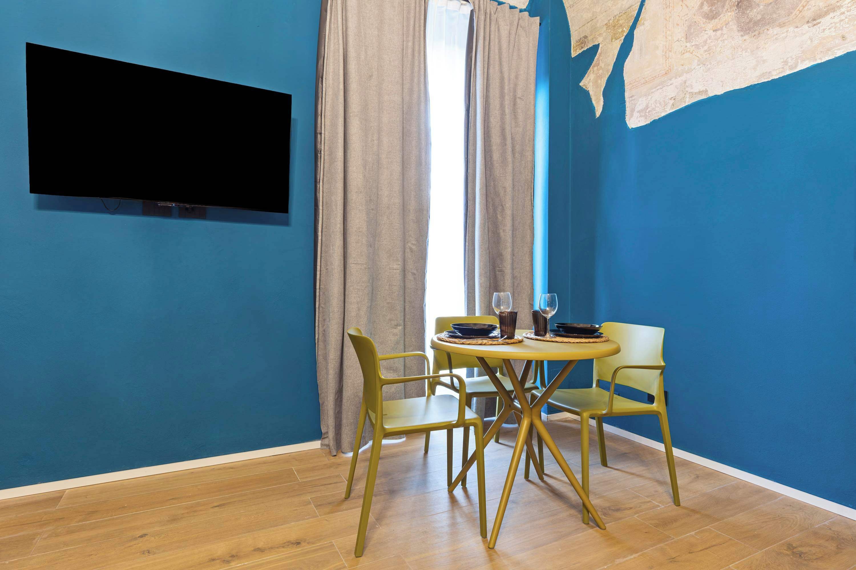Une pièce avec un mur bleu, un téléviseur à écran plat, une table à manger avec des chaises et une fenêtre avec des rideaux.