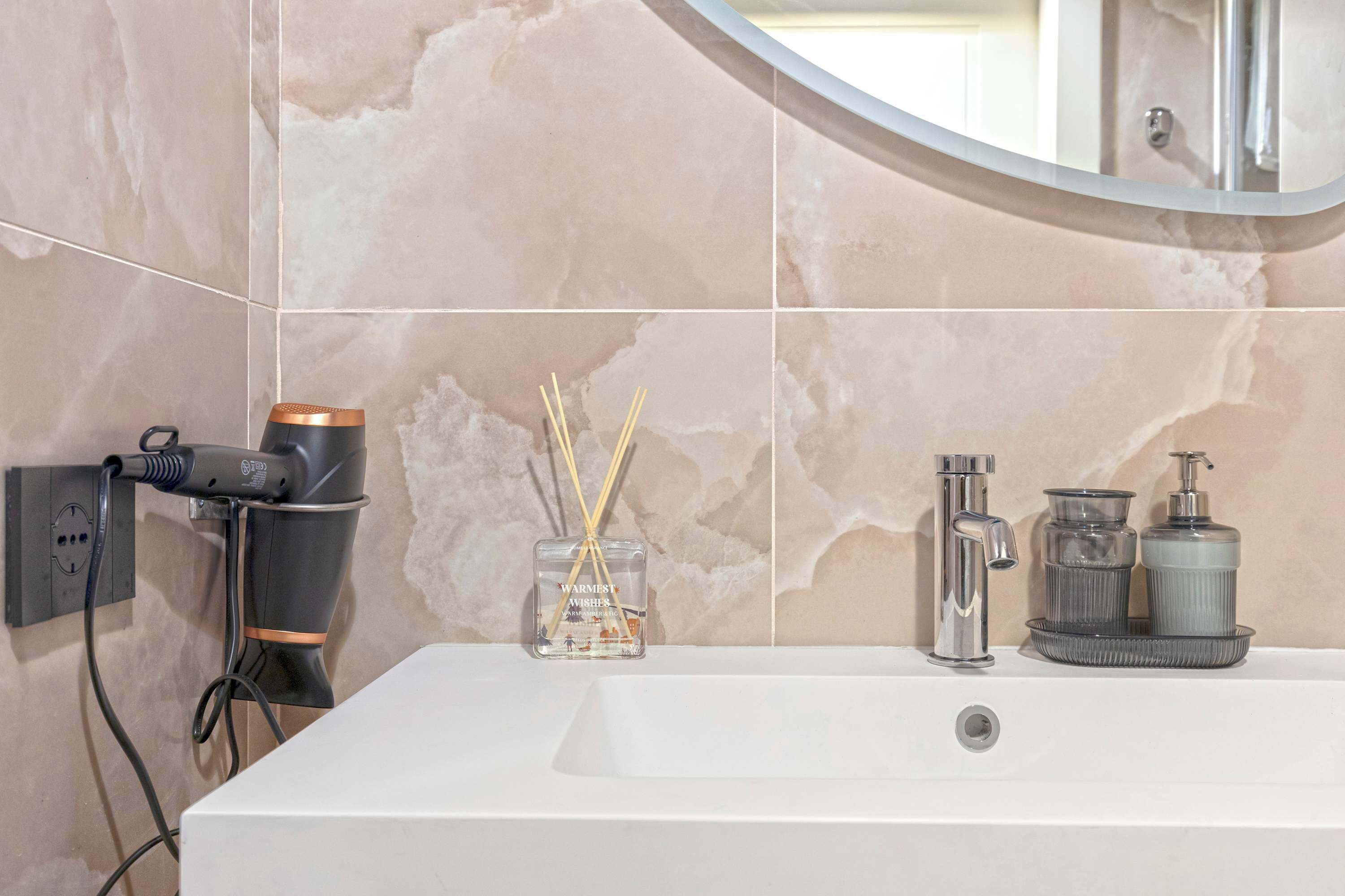 A bathroom with a white sink, a mirror, a hairdryer, and a small vase with reeds.