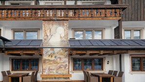 A building with a wooden balcony, large windows, and a central mural. - ALPRESORT Hubertushof (Oberstaufen)
