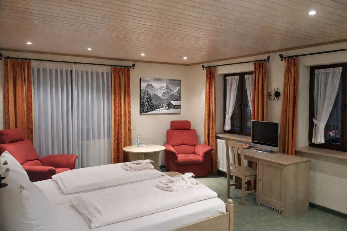 A hotel room with a bed, two red armchairs, a small table with a bottle, a desk with a computer, and a window with curtains.