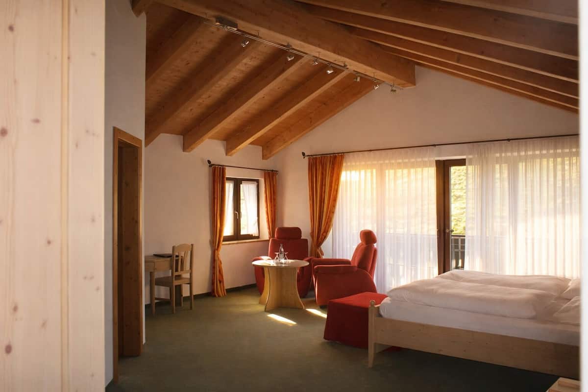 A room with a bed, two red chairs, a small table, and a window with curtains.