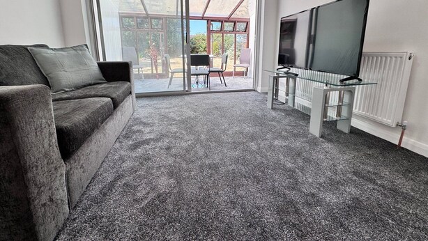 Ein Wohnzimmer mit grauem Sofa, einem Glastisch, einem Flachbildfernseher und einer Schiebetür zu einer Außenpatio mit Sitzgelegenheiten.