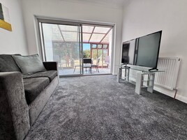 Una sala de estar con un sofá gris, una mesa de centro de vidrio, un televisor de pantalla plana y un cerramiento de vidrio corredizo que da a un patio exterior con sillas.