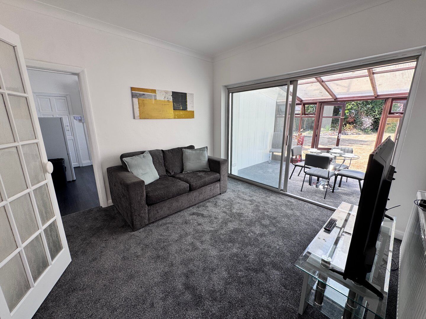 A modern living room with a grey sofa, a flat-screen TV, and a sliding glass door leading to an outdoor patio area.
