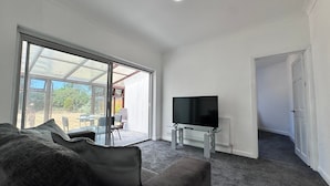 Ein modernes Wohnzimmer mit einer großen Schiebeglasdoor zur Außenbereits, einem an der Wand montierten Flachbildfernseher und einem dunkelgrauen Sofa.