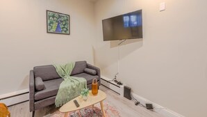 Un salon avec un canapé gris, une petite table basse ronde, une télécommande, une plante et un téléviseur fixé au mur.