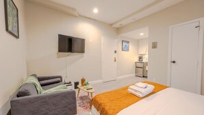 Une chambre d’hôtel avec un lit, un canapé, une télévision, une petite table avec une télécommande, et un coin cuisine à l’arrière-plan.