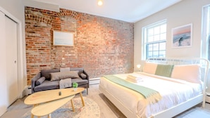 A bedroom with a brick wall, a bed, a sofa, and a small table. - Peaceful South End Studio! (Boston)