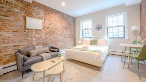 A modern bedroom with a brick wall, a bed, a sofa, a small table, and a chair. - Peaceful South End Studio! (Boston)