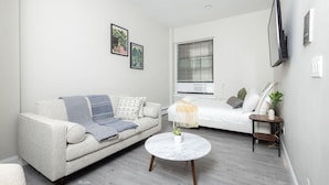 Sala de estar moderna e minimalista com um sofá cinza, uma mesa de centro redonda e uma cama com edredom branco. Há duas molduras na parede, uma janela com persianas e uma televisão na parede.