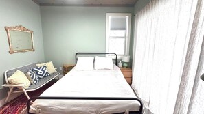 A small, simple bedroom with a single bed, a chair, a nightstand, a lamp, and a window with curtains. - Cute 2-bedroom house in Jay, perfect for a relaxing getaway. (Jay)