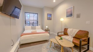 A bedroom with a bed, a TV mounted on the wall, a window with blinds, a bedside table with a lamp, a sofa, and a small rug. - Peaceful Studio | Close to Fenway (Boston)