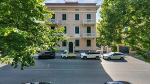 Ein mehrstöckiges Gebäude mit einem zentralen Eingang, einem Parkplatz mit mehreren Autos und Bäumen, die den Blick einfassen.