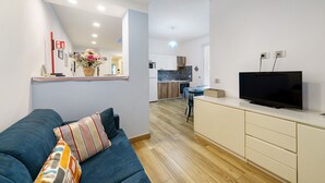 Ein Wohnzimmer mit einer Couch, einem Fernseher auf einem Schrank, einer Küche im Hintergrund und einem kleinen Essbereich.