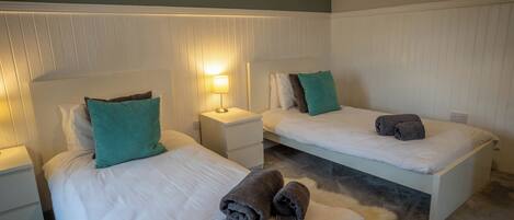 A hotel room with two single beds, white bedding, teal pillows, and a small bedside table with a lamp.