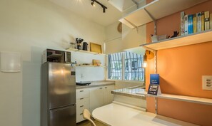A compact kitchen with a stainless steel refrigerator, a microwave, and a small countertop. There are shelves with books and a window providing natural light. - Fully Equipped Studio in Asa Norte Edl0106 (Brasília)