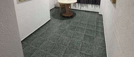 A small, narrow room with a tiled floor, a round table, a chair, and a window with curtains.
