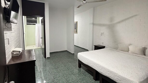 Une chambre à coucher comprenant un lit, un ventilateur de plafond, une table de chevet, une commode et une salle de bain visible à travers une porte ouverte.