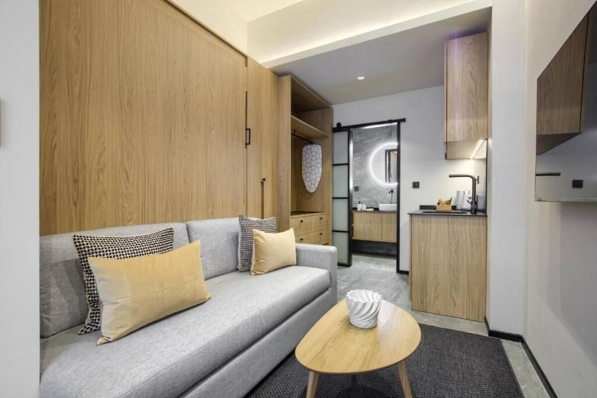 Une chambre d’hôtel moderne avec un canapé gris, des meubles en bois, une petite table ronde et un coin cuisine équipé d’un évier et d’un plan de travail.