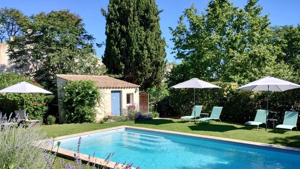 A rectangular swimming pool with a tiled edge, surrounded by a grassy area with lavender plants. There are two umbrellas and two lounge chairs in the background. - Le Petit Molière Gîte for 2 people (Servian)