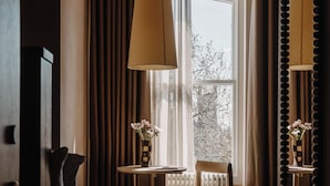 A hotel room with a bed, a table with a chair, a window with curtains, a radiator, and a mirror with a decorative frame. - Mason and Fifth - Belsize Park (London)