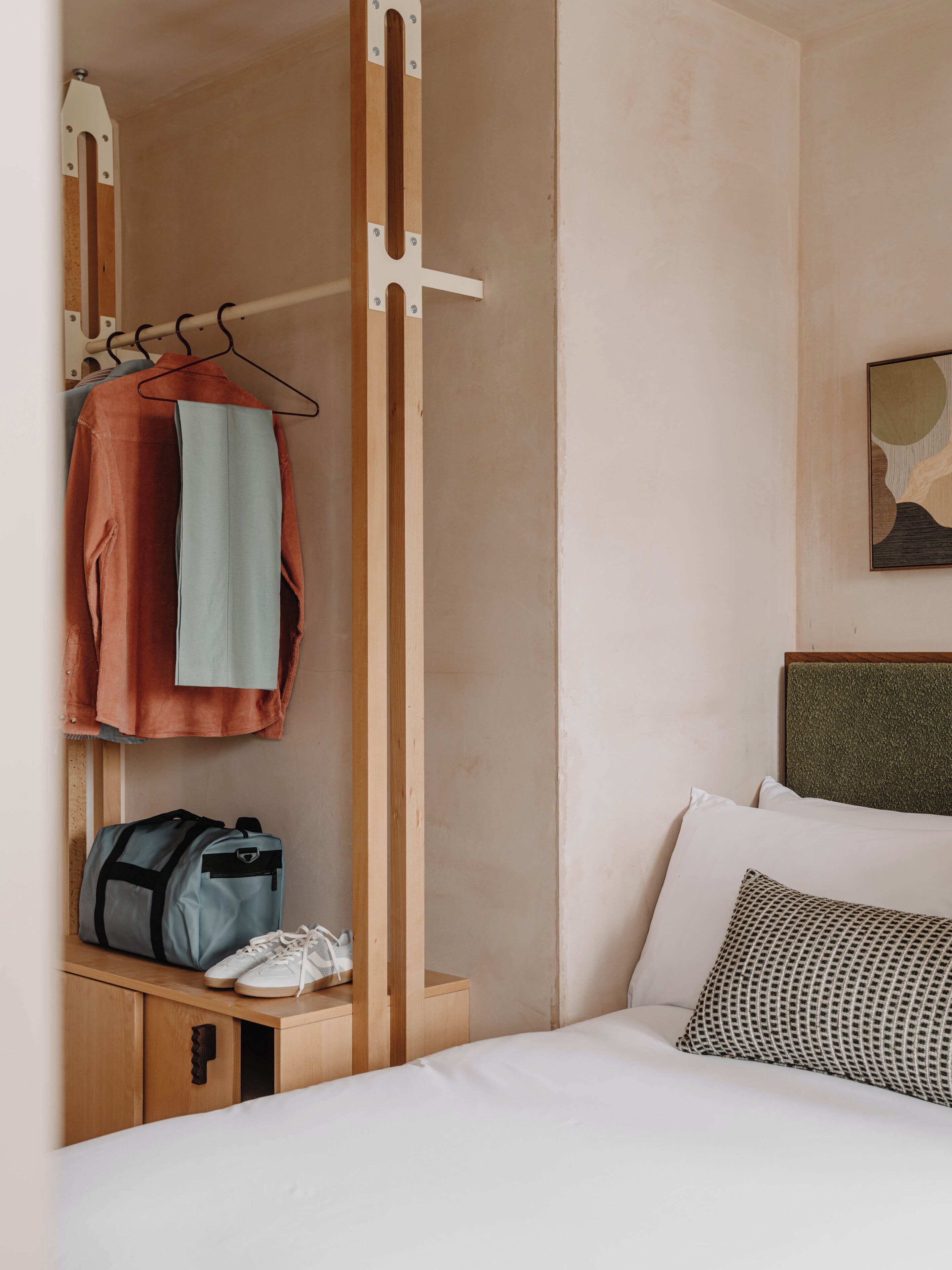 Quarto de hotel com uma cama, um cabide suspenso, uma pequena mesa de cabeceira de madeira e uma obra de arte na parede.