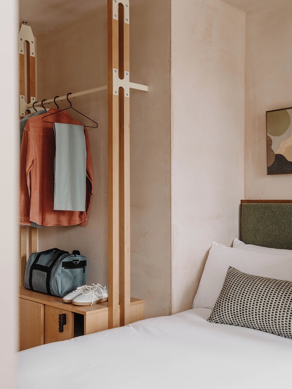Camera d'albergo con un letto, un appendiabiti, un comodino in legno e un'opera d'arte appesa al muro.