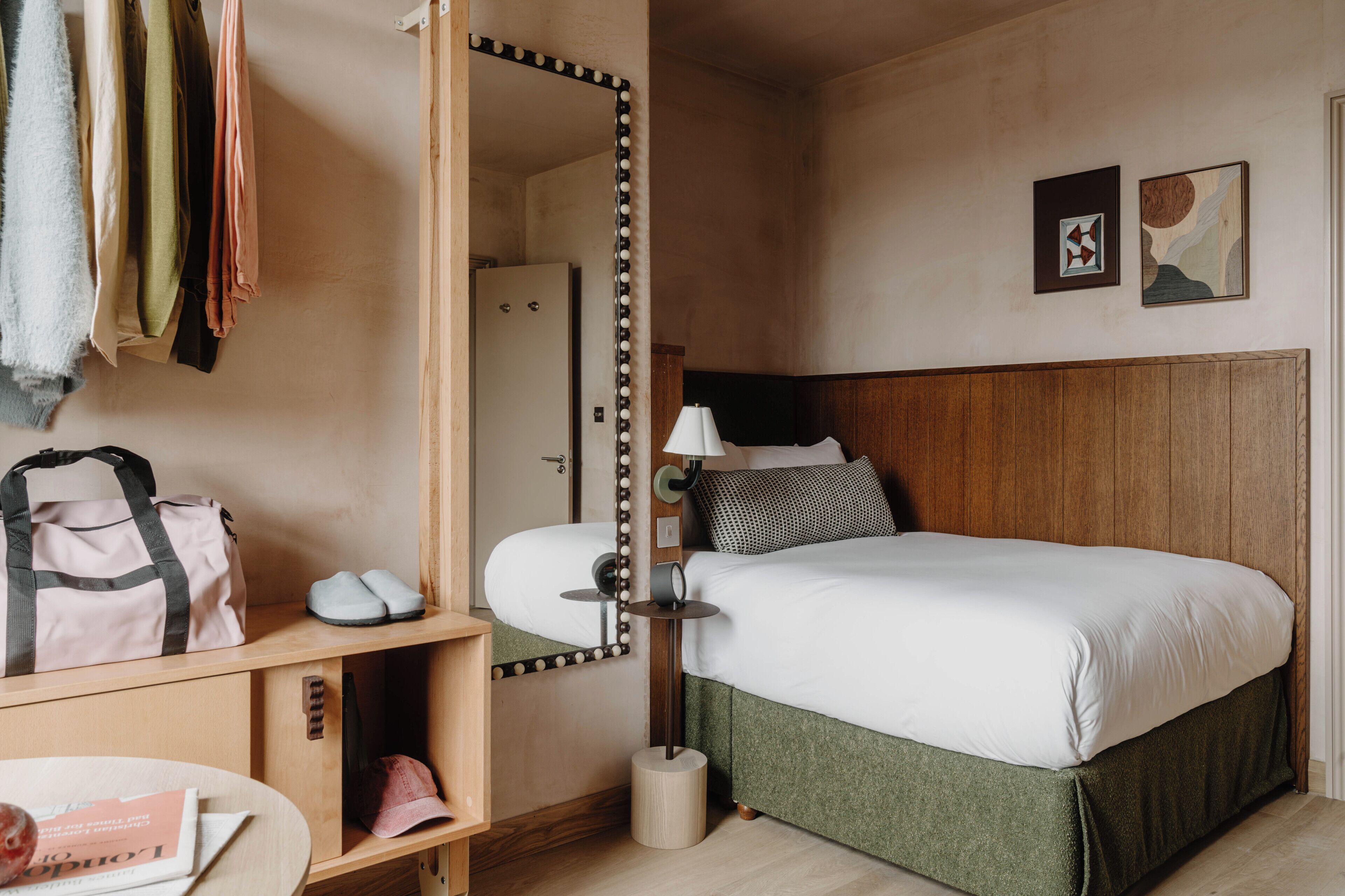 Ein Hotelzimmer mit Bett, Spiegel, Lampe, Schuhablage und Kleiderhaken mit Kleidung.