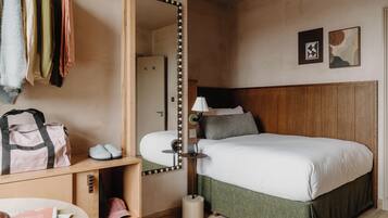 Ein Hotelzimmer mit Bett, Spiegel, Lampe, Schuhablage und Kleiderhaken mit Kleidung.