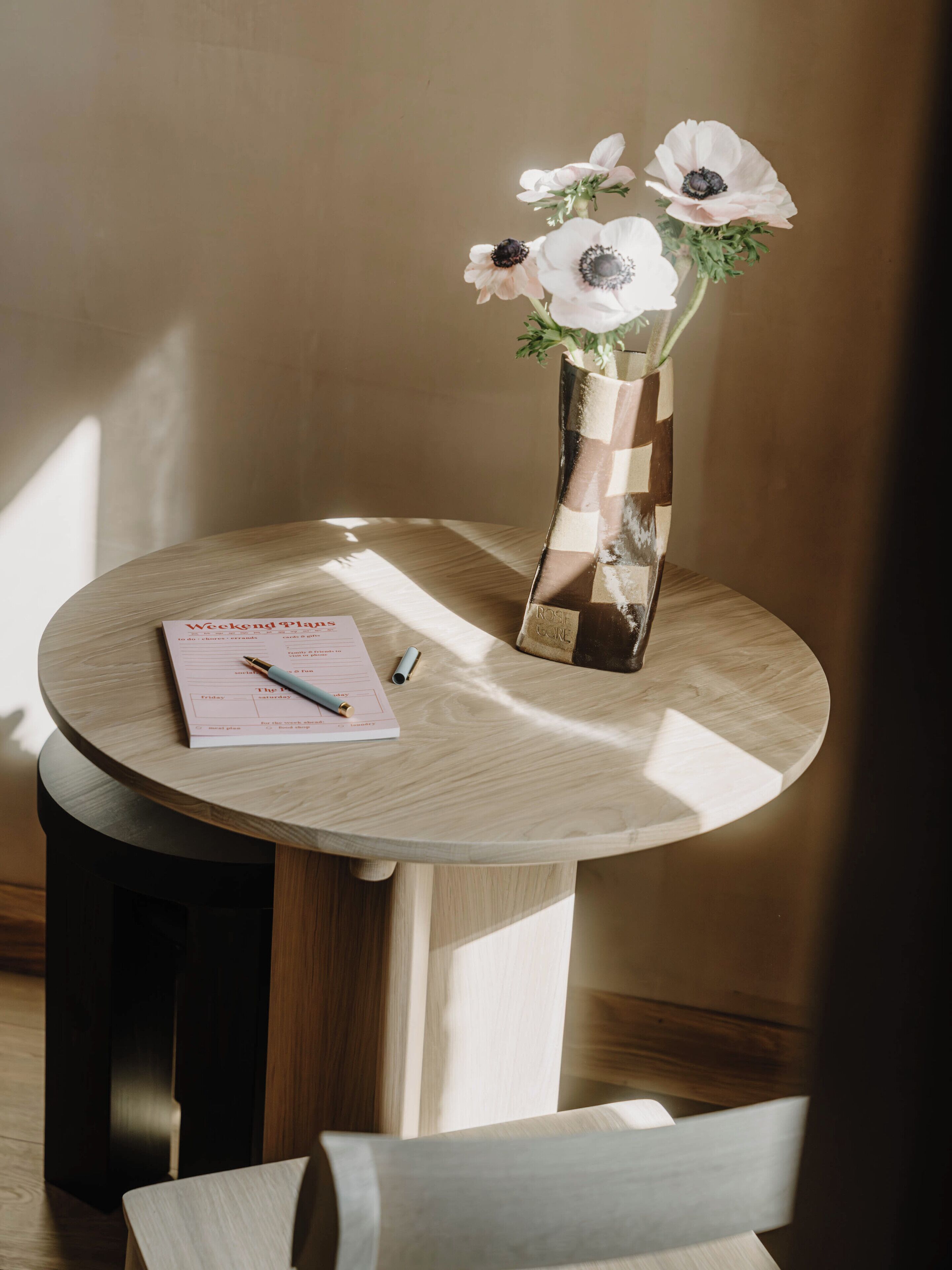 A round wooden table with a checkered vase containing white flowers, a notepad, and a pen.