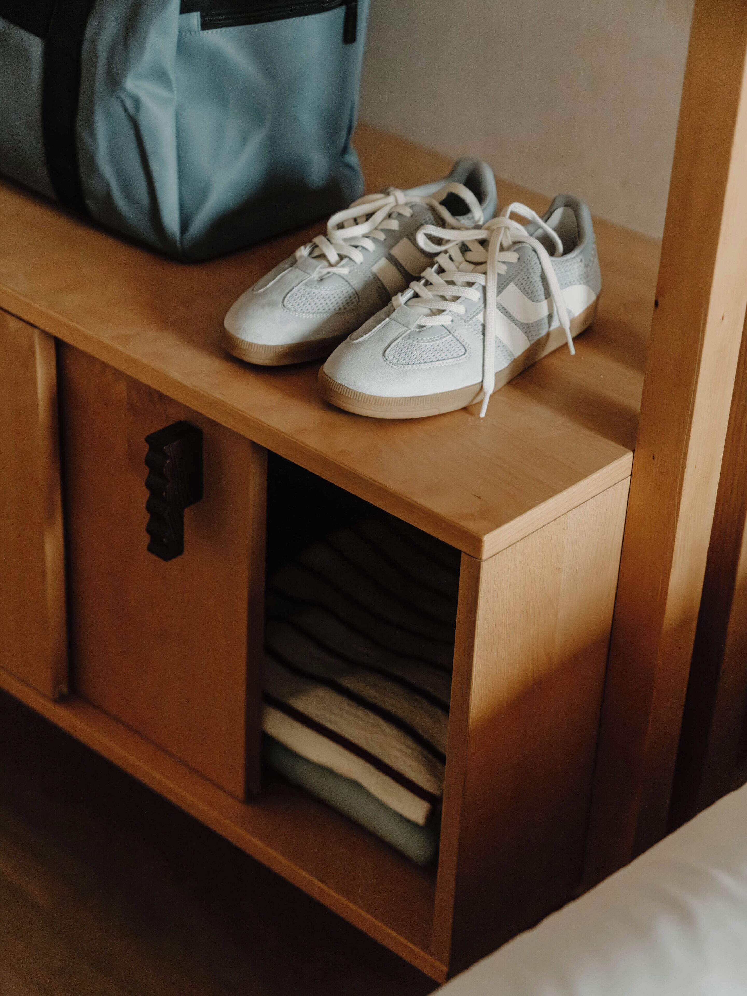 Una mesita de noche de madera con un par de zapatillas blancas encima y un bolso azul a su lado.